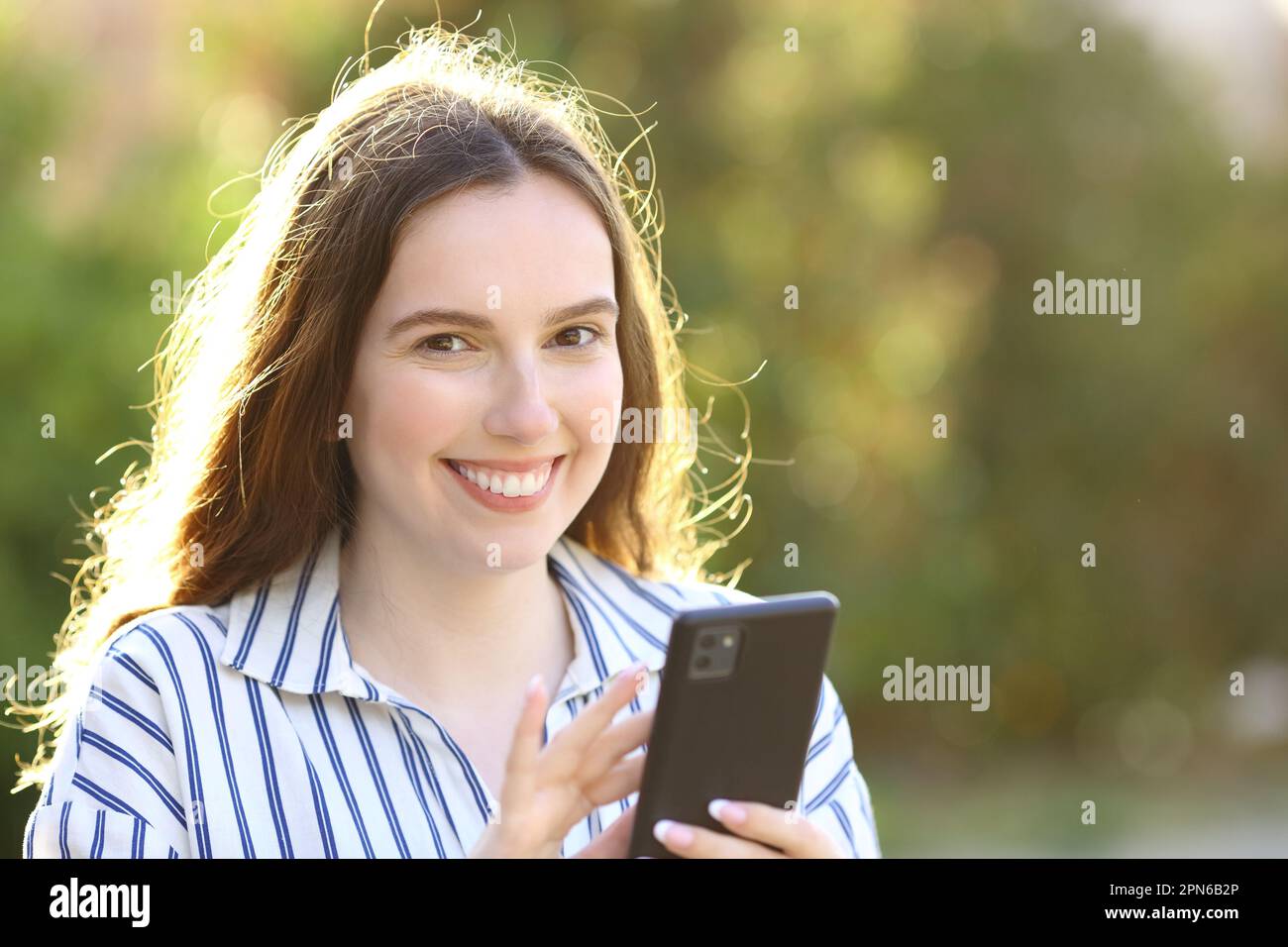 Glückliche Frau mit Handy sieht in die Kamera, wenn sie in einem Park steht Stockfoto