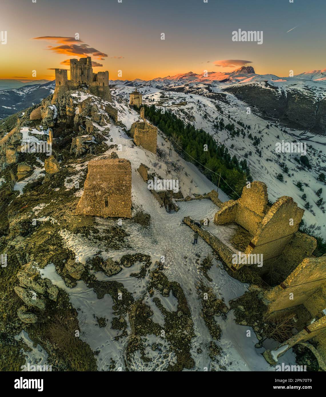 Blick aus der Vogelperspektive auf die Rocca di Calascio mit Schnee und im Licht des Sonnenuntergangs beleuchtet, hinter der Kette der Gran Sasso und des Corno grande. Stockfoto