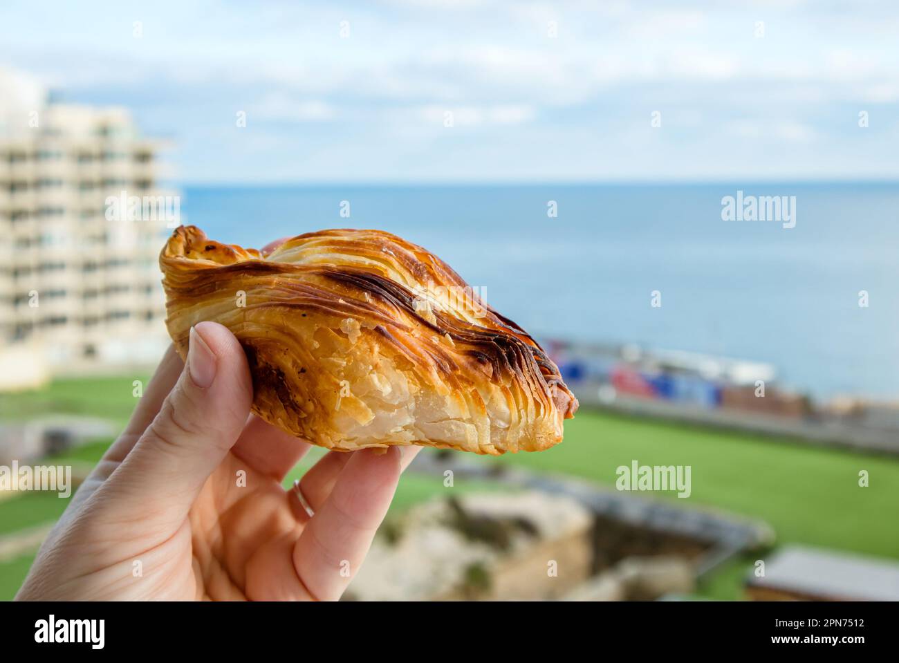 Lokales maltesisches Essen namens Pastizz oder Pastizzi. Person hält rautenförmigen kleinen Kuchen mit Malta Sliema Stadt auf dem Hintergrund mit blauem Himmel und Meer Stockfoto