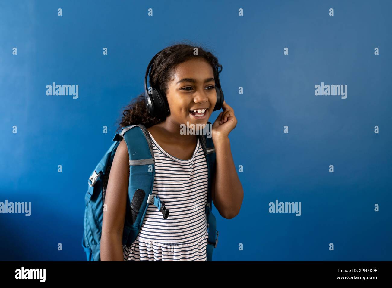 Fröhliches, birassisches Schulmädchen mit Kopfhörern, das Musik auf blauem Hintergrund mit Kopierbereich hört Stockfoto