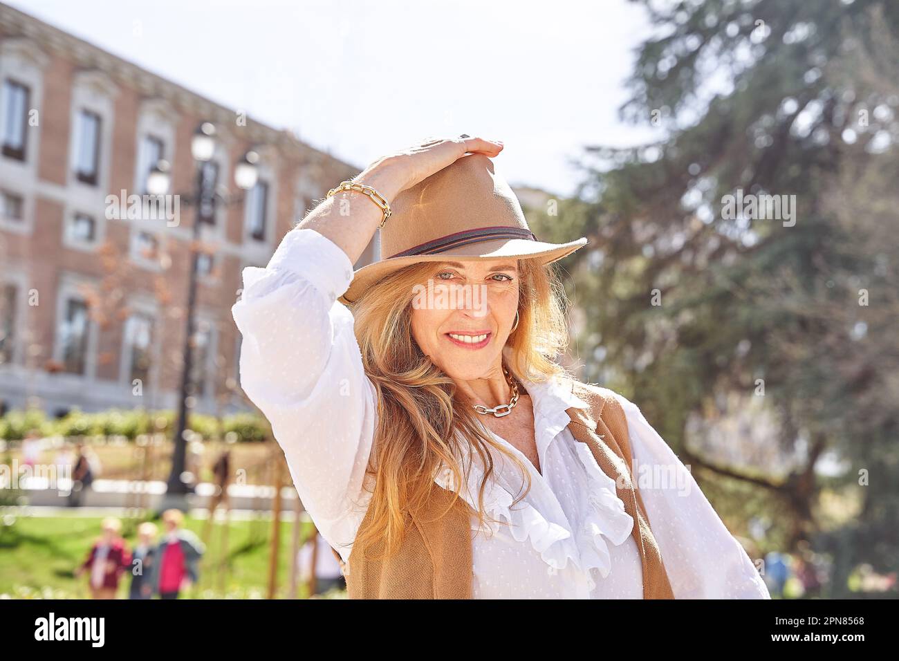 Eine sorgenfreie blonde Frau mit langem Haar und einem stilvollen Hut blickt direkt in die Kamera und genießt die friedliche Umgebung eines Parks. Stockfoto