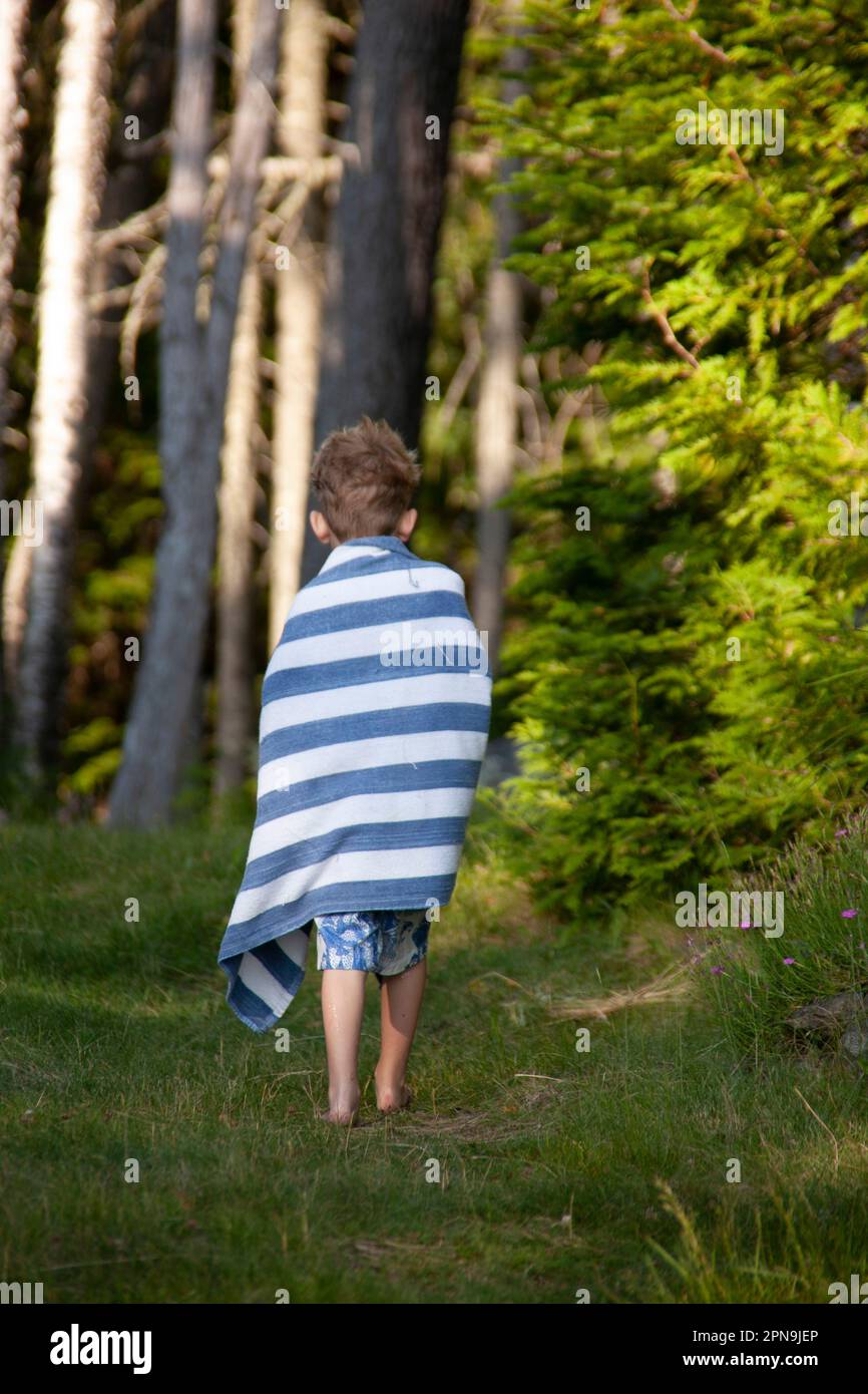 Rückansicht eines kleinen Jungen, der auf einem grasbedeckten Pfad mit gestreiftem Handtuch spaziert Stockfoto