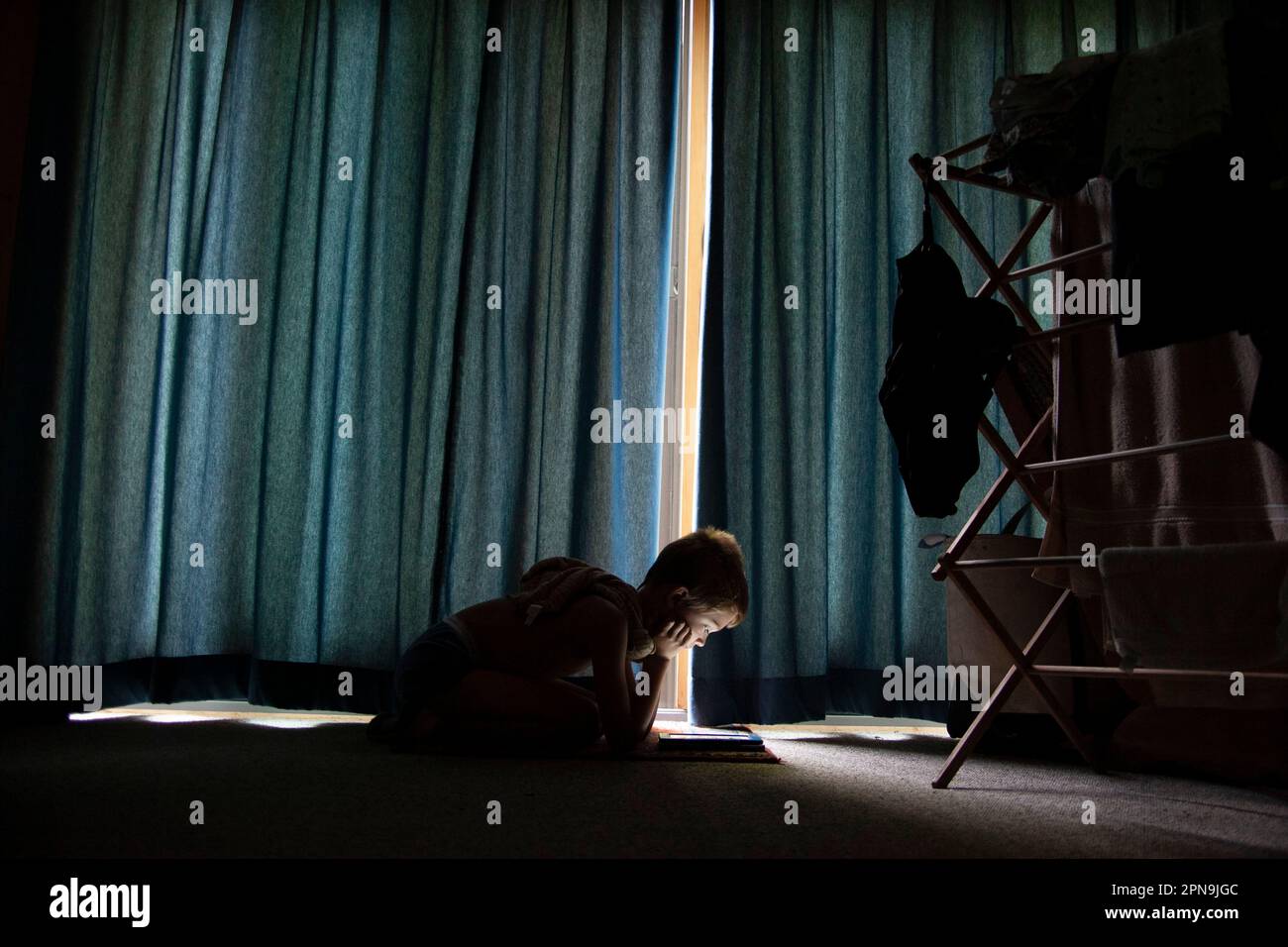Ein kleiner Junge auf dem Boden, der liest, wenn das Sonnenlicht durch geschlossene Vorhänge sickert Stockfoto