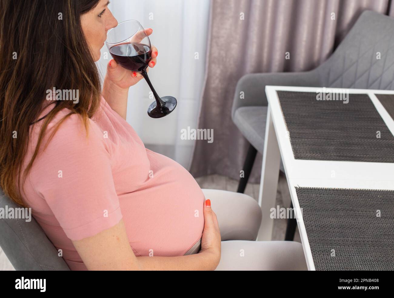 Ein schwangeres Mädchen sitzt in der Küche am Tisch und trinkt Alkohol. Schlechte Angewohnheit während der Schwangerschaft. Stockfoto