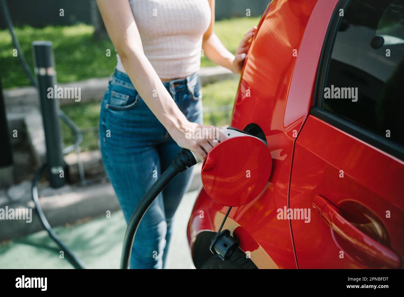 Schneide eine Frau in lässigem Outfit, indem du das Ladegerät in das rote Auto steckst, während du in der EV-Ladestation stehst Stockfoto
