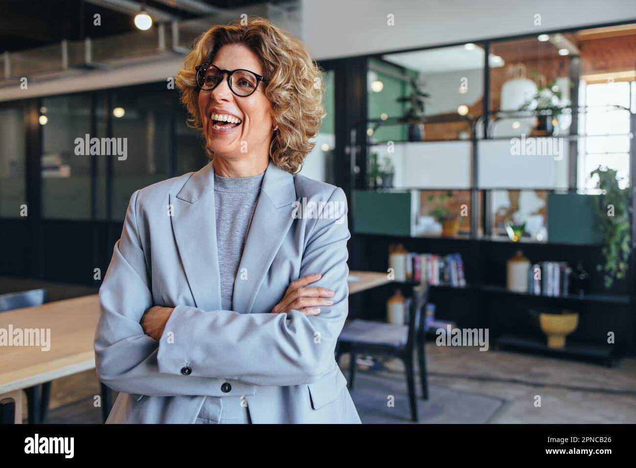 Reife Geschäftsfrau, die lächelt und mit gekreuzten Armen steht. Eine Geschäftsfrau, die einen Anzug im Büro trägt. Stockfoto