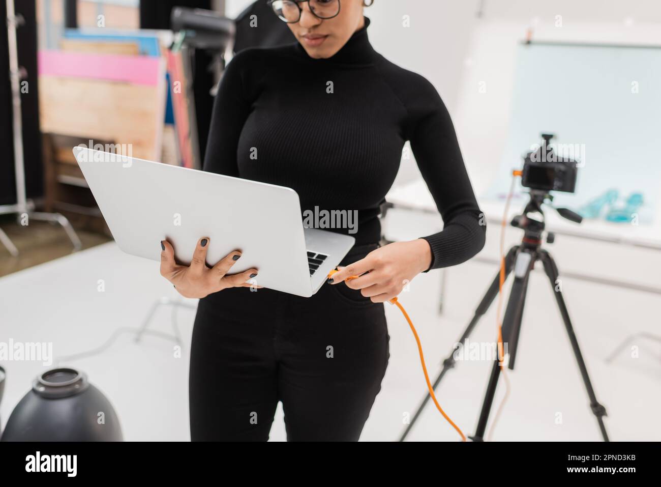 afroamerikanischer Content Produzent, der ein Kabel mit einem Laptop in der Nähe einer verschwommenen Digitalkamera im Fotostudio verbindet, Stock Image Stockfoto