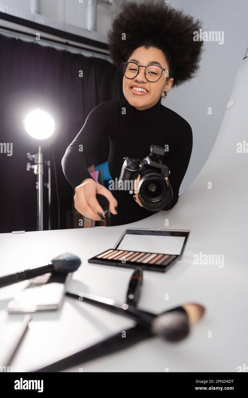 Fröhlicher afroamerikanischer Fotograf mit Brille, der in der Nähe dekorativer Kosmetika auf dem Schießtisch im Fotostudio auf die Kamera schaut, Stockbild Stockfoto