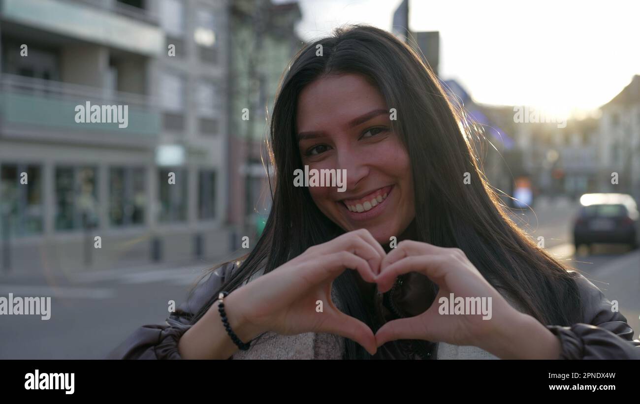 Eine glückliche junge Frau, die ein Herzsymbol trägt, mit Händen, die während des Sonnenuntergangs auf der Straße der Stadt stehen. Das Porträtgesicht einer Person macht eine herzförmige Geste Stockfoto
