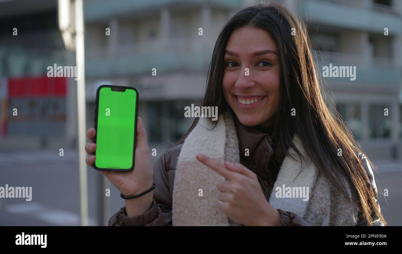 Eine fröhliche junge Frau zeigte ihr Handy vor der Kamera mit einem grünen Bildschirm mit Chroma Key auf der Straße der Stadt. Weibliche Person, die mit dem Finger auf den Bildschirm des Mobiltelefons zeigt V Stockfoto
