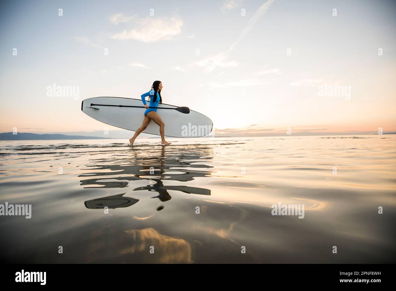 Eine Frau, die bei Sonnenuntergang am Strand mit einem Paddleboard rennt Stockfoto