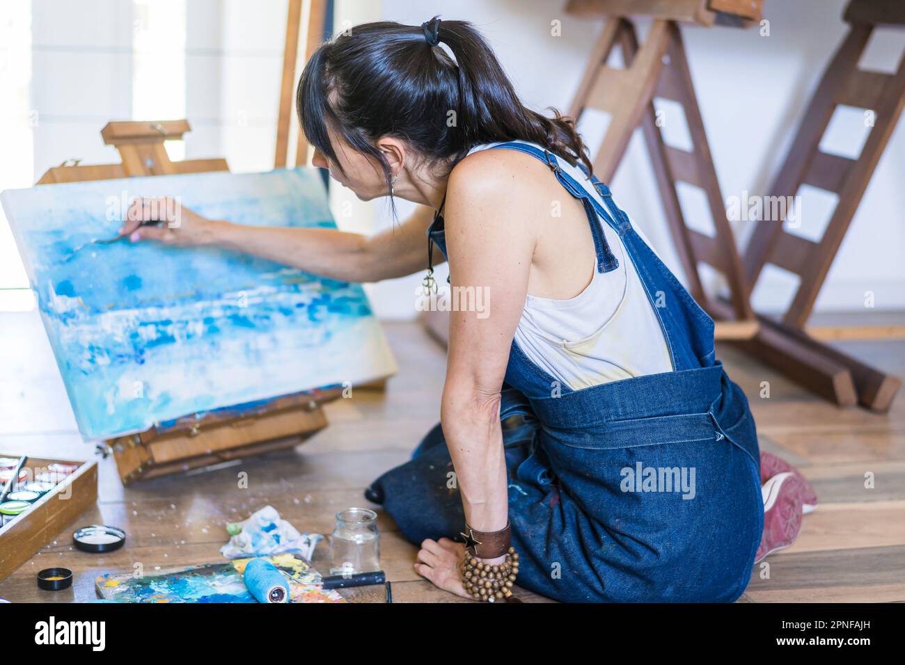 Künstlerin malt auf einer Leinwand ein blaues abstraktes Gemälde. Kreative Frau, die in ihrem Kunstatelier auf dem Boden arbeitet. Stockfoto