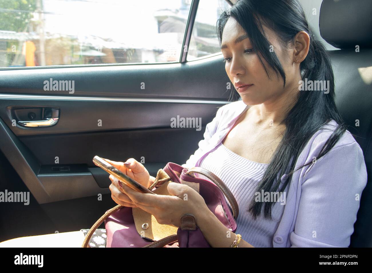 Eine junge Frau mit Handtasche beobachtet ihr Handy, während sie im fahrenden Auto sitzt Stockfoto