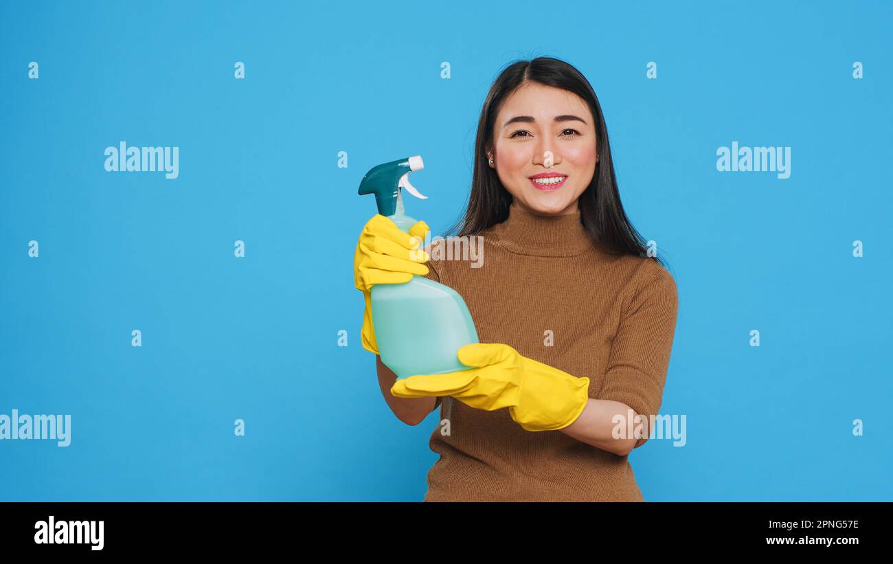 Lächelndes asiatisches Zimmermädchen trägt Schutzhandschuhe und zeigt chemisches Reinigungsspray vor der Kamera, um das Reinigungsprodukt zu fördern. Professionelle Haushälterin hat hart gearbeitet, um ein sauberes Zuhause zu erhalten Stockfoto