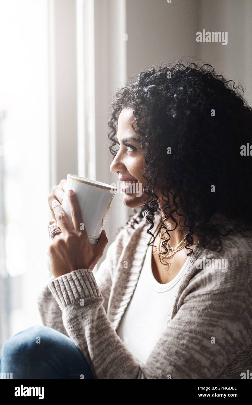 Wie glücklich ich bin, zu Hause zu bleiben. Eine fröhliche junge Frau, die tagsüber eine Tasse Kaffee genießt, während sie zu Hause auf einem Stuhl sitzt. Stockfoto