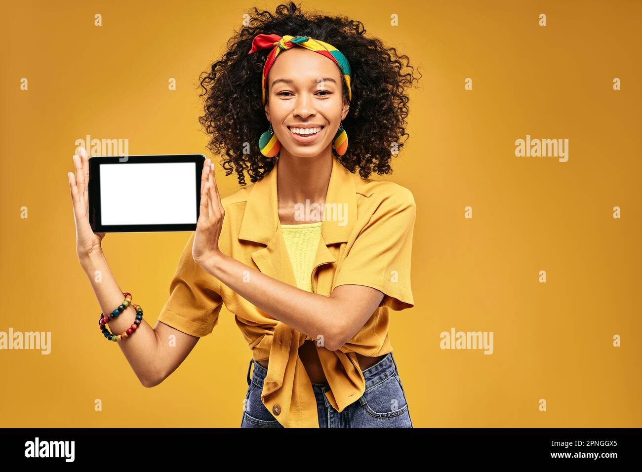 Eine Rastafarin in fröhlicher, leuchtender Kleidung mit einem digitalen Tablet in den Händen, um Hipster-Produkte auf gelbem Hintergrund zu bewerben Stockfoto