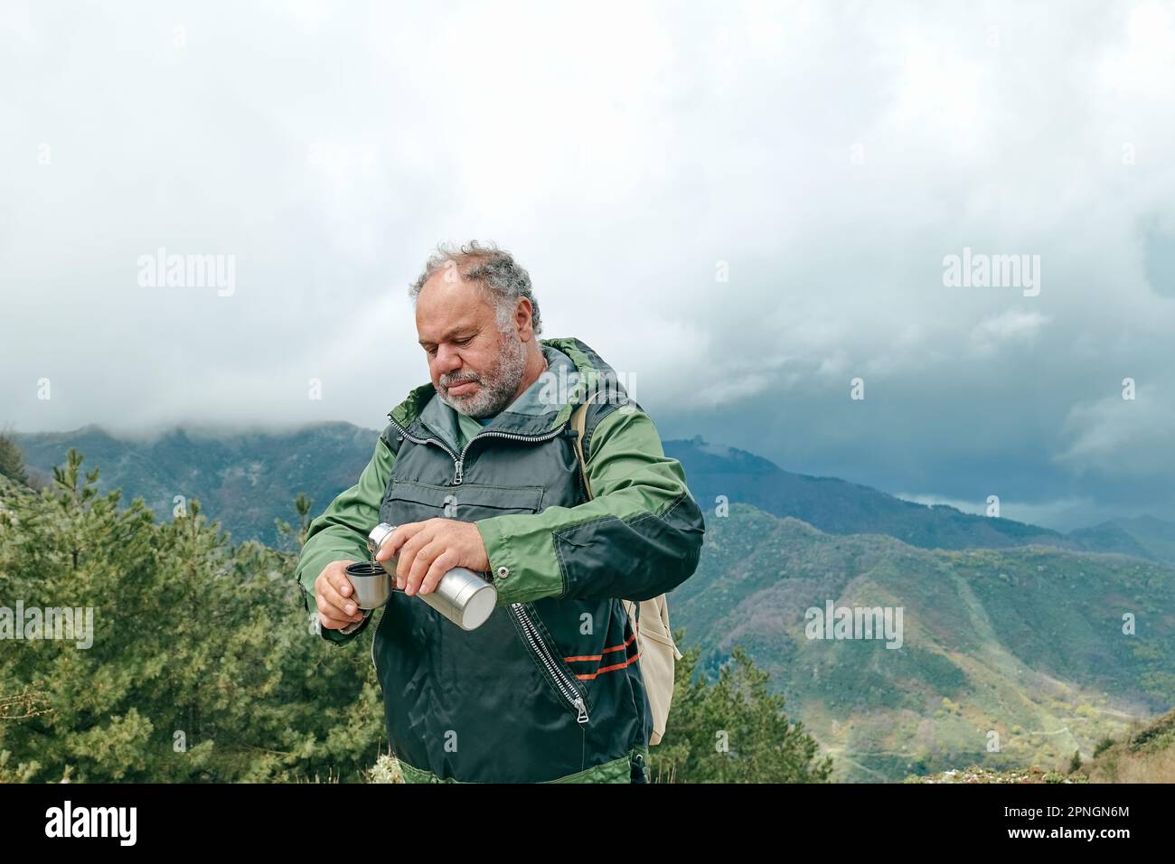 Reisender Mann wandert im Bergwald, entspannt sich, während er eine Tasse Kaffee aus der Thermoskanne im Panorama-Bergtal trinkt. Stockfoto