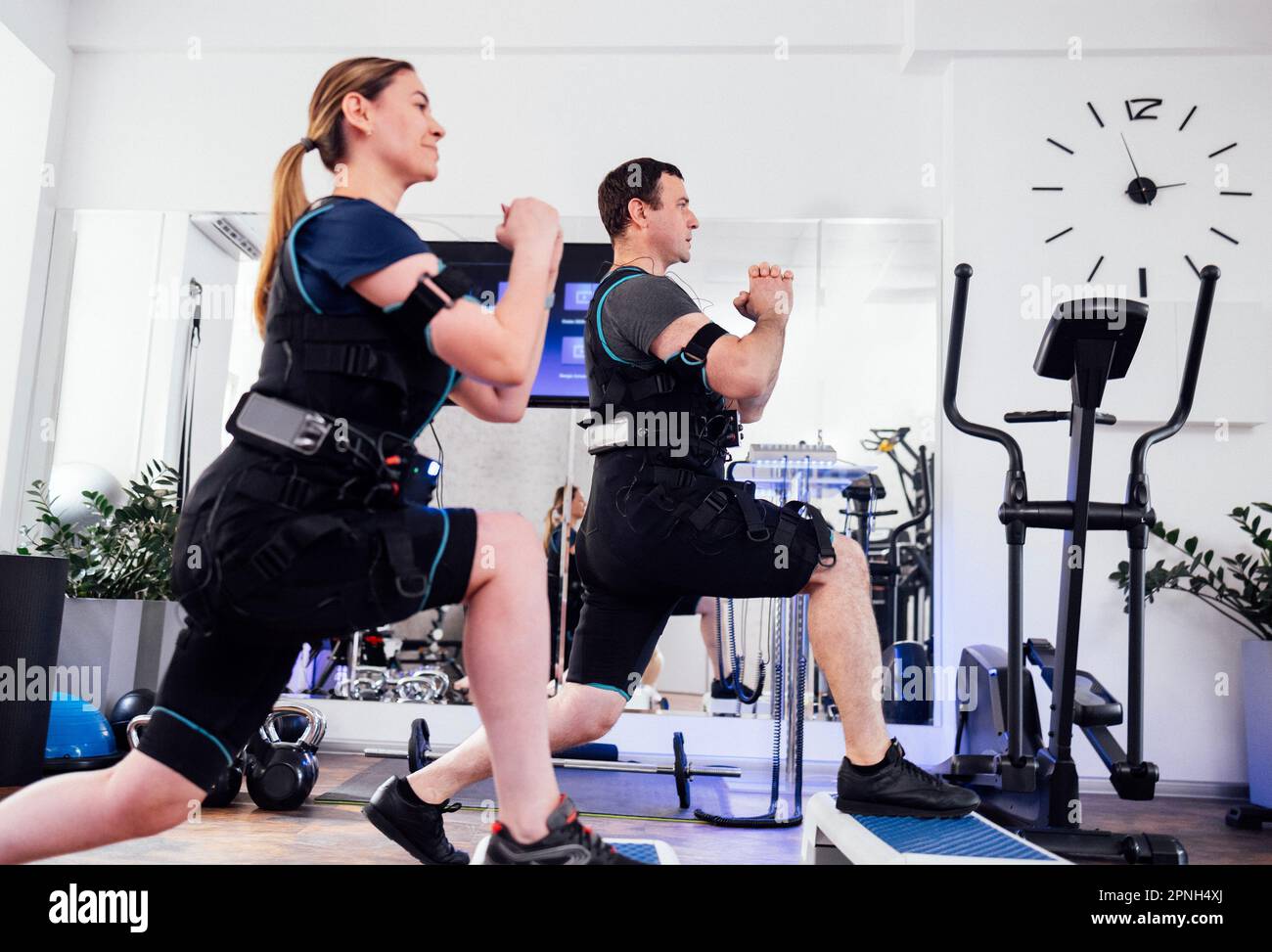 Sportpaar in ems-Anzügen ist körperlich im Fitnessclub oder Fitnessraum aktiv. Mann und Frau mit geballten Händen, die mit Step-pla nach vorne Ausfallschritte machen Stockfoto