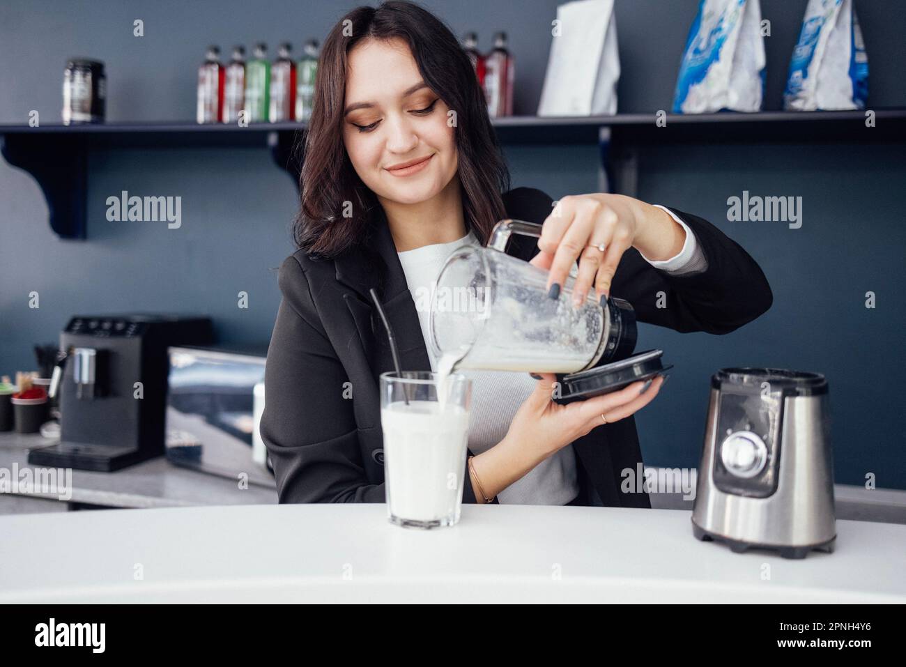 Junge hübsche Frau bereitet Proteinshake mit stationärem Mixer zu. Die Barista gießt Milchshake mit Strohhalm in das Glas. Kaffeemaschine, Mikrowelle Stockfoto