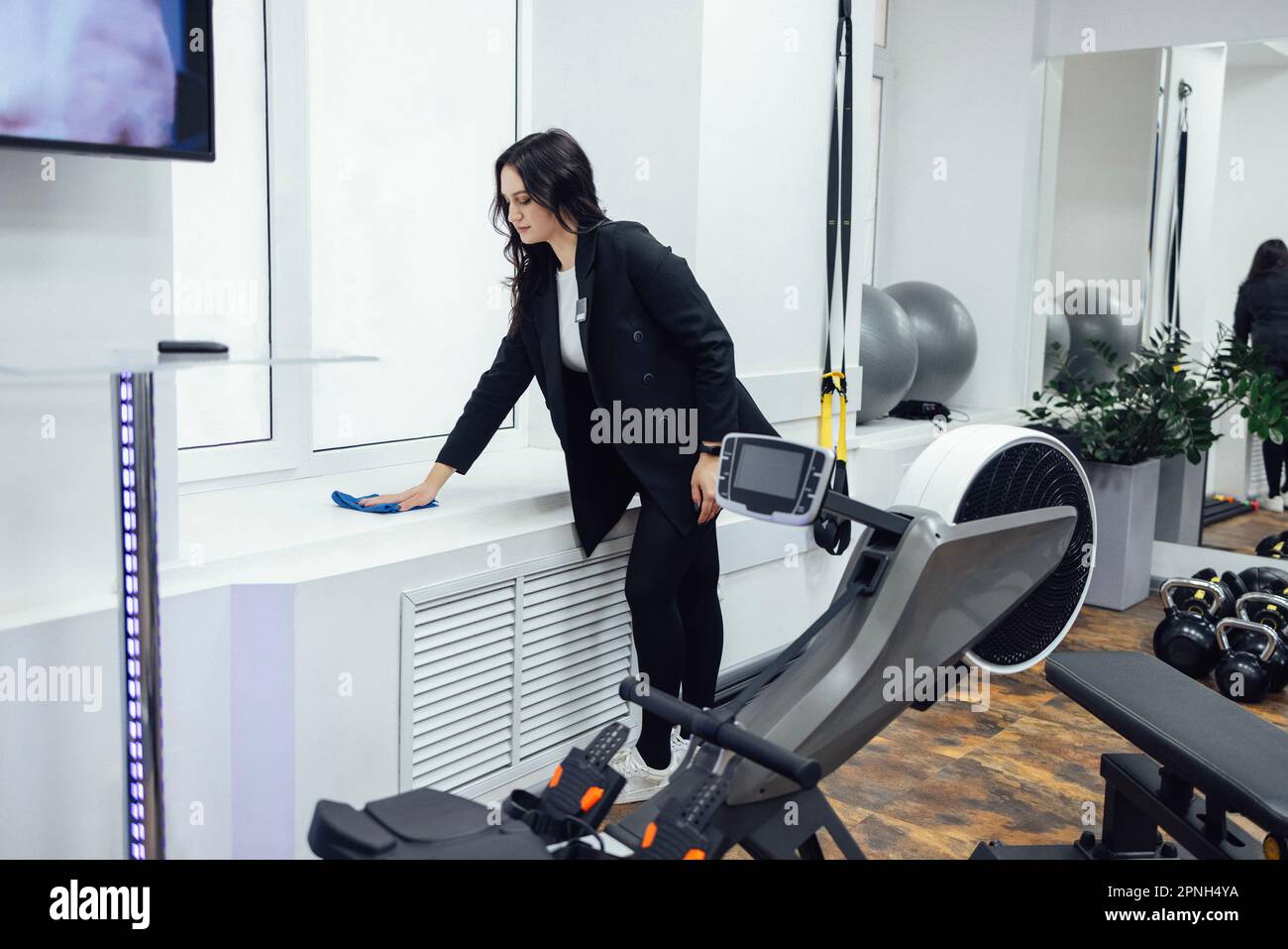 Junge brünette Frau in schwarzer Jacke wischt Fensterbank im Fitnessclub. Das Personal räumt im Fitnessraum auf. Moderne, elegante Einrichtung in hellen Farben mit Sportausstattung Stockfoto