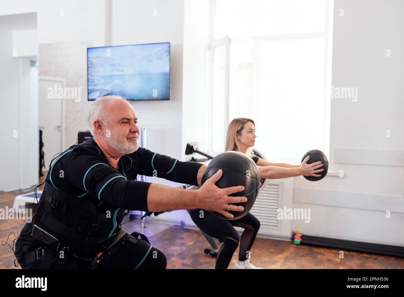 Der grauhaarige ältere Mann im ems-Anzug trainiert mit einem persönlichen Fitnesstrainer in einem modernen Fitnessraum. Alte männliche Sportlerbesetzungen mit gewichtetem Ball bei Functional e Stockfoto