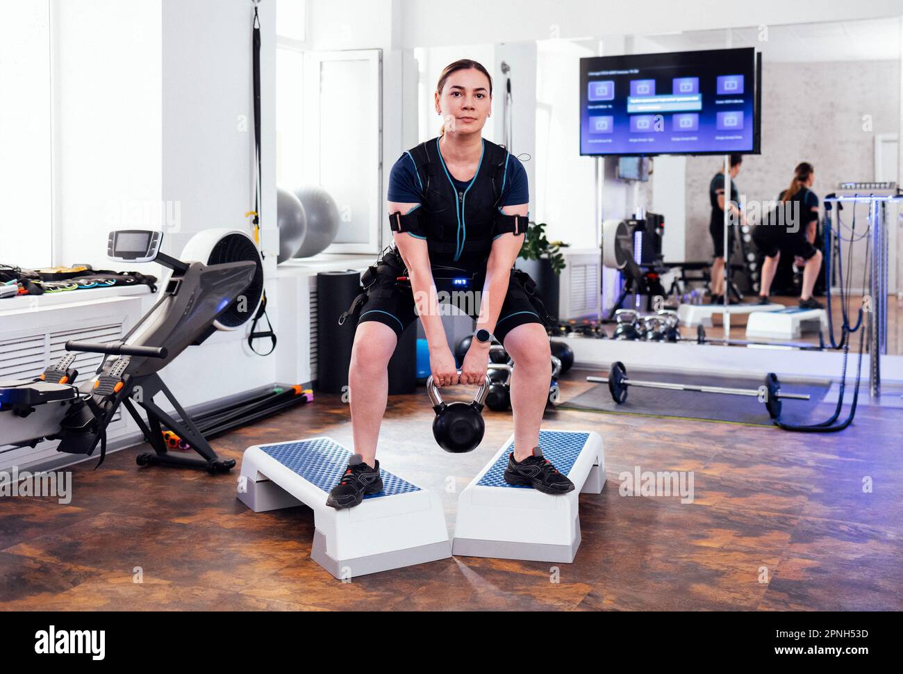 Junge Frau im rettungssanitäter-Anzug steht auf Treppen und macht Squats mit Kugelhantel. Weibliche Athleten, die mit Gewicht in einem modernen Fitnessstudio hocken. Elektrischer Muskelstich Stockfoto