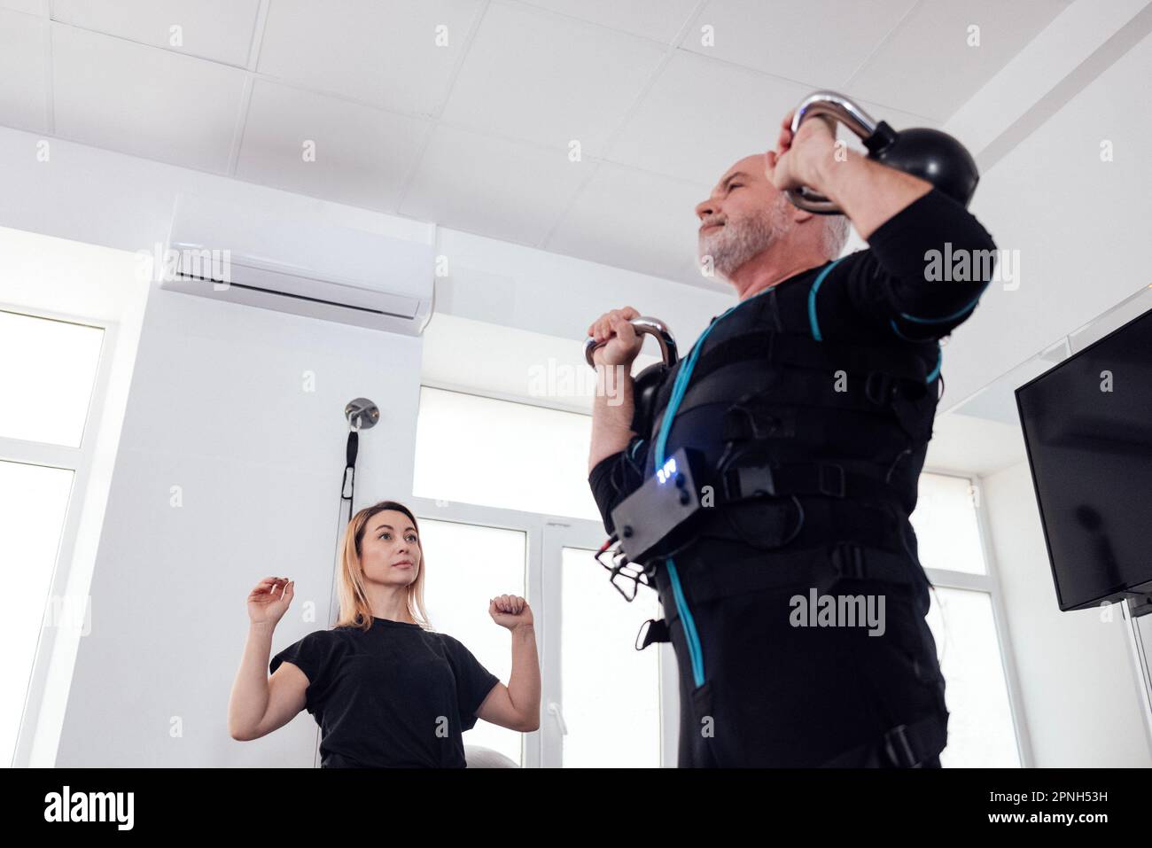 Der grauhaarige ältere Mann im ems-Anzug trainiert mit einem persönlichen Fitnesstrainer in einem modernen Fitnessraum. Alter Sportler mit Kugelhanteln im funktionellen Stockfoto