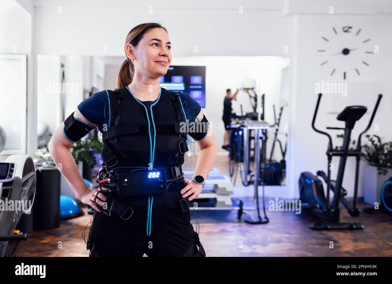 Junge lächelnde Frau, die sich in einem elektrischen Muskelstimulationskostüm im Fitnessclub oder Gym posiert. Selbstbewusste Sportlerin im rettungssanitäter-Anzug mit den Händen an ihrem Bell Stockfoto