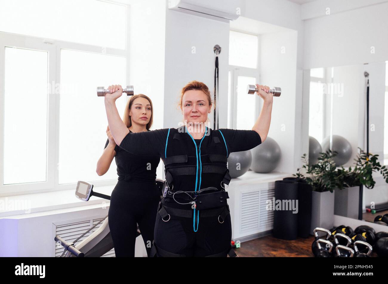 Eine Frau mittleren Alters im Anzug zur elektrischen Muskelstimulation ist mit einem persönlichen Trainer an körperlichen Aktivitäten beteiligt. Eine dicke Frau, die mit der Kurzhantel trainiert Stockfoto