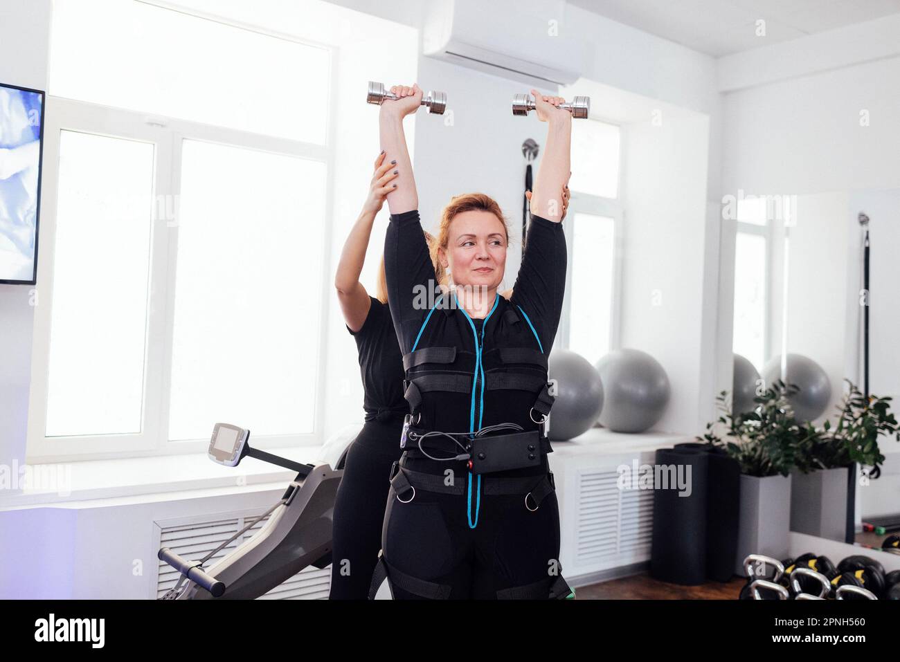 Eine lächelnde Sportlerin mittleren Alters im rettungssanitäter-Anzug ist mit einem persönlichen Trainer körperlich aktiv. Blonde, dicke Frau, die ihre Hände mit Kurzhantel hebt Stockfoto