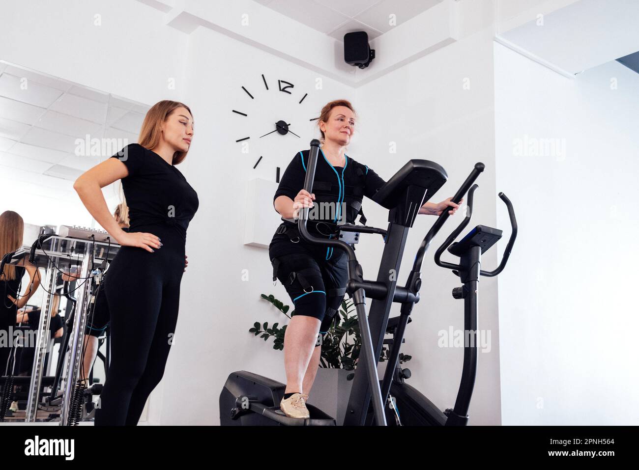 Ältere Frau im Anzug zur elektrischen Muskelstimulation trainiert mit ihrem persönlichen Trainer im Fitnessstudio oder Fitnessstudio auf einem Ellipsentrainer. Junge weibliche t Stockfoto