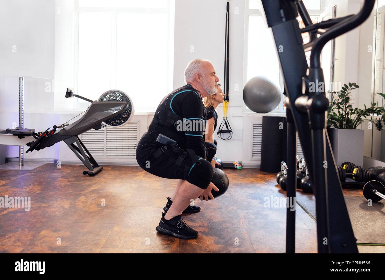 Ein älterer Mann im rettungssanitäter-Anzug trainiert mit einem persönlichen Trainer im Fitnessstudio oder Fitnessstudio. Alte Sportlerbesetzungen mit gewichtetem Ball bei elektrischer Muskelreizung Stockfoto