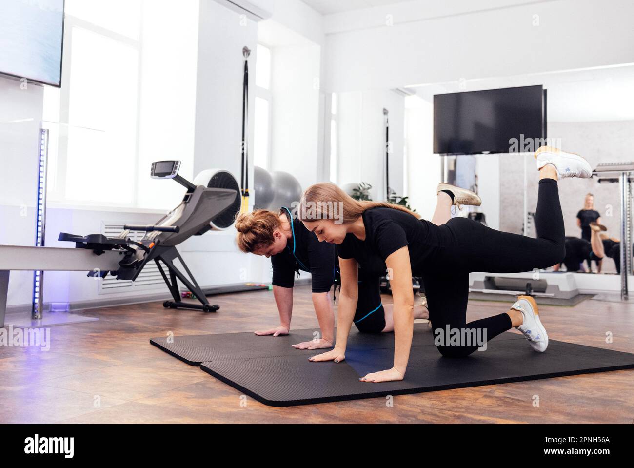 Eine blonde Frau mittleren Alters im rettungssanitäter-Anzug trainiert mit einem Personal Trainer im Fitnessstudio. Frau, die Übungen macht, um die Muskeln von Rücken und Hüften zu stärken Stockfoto