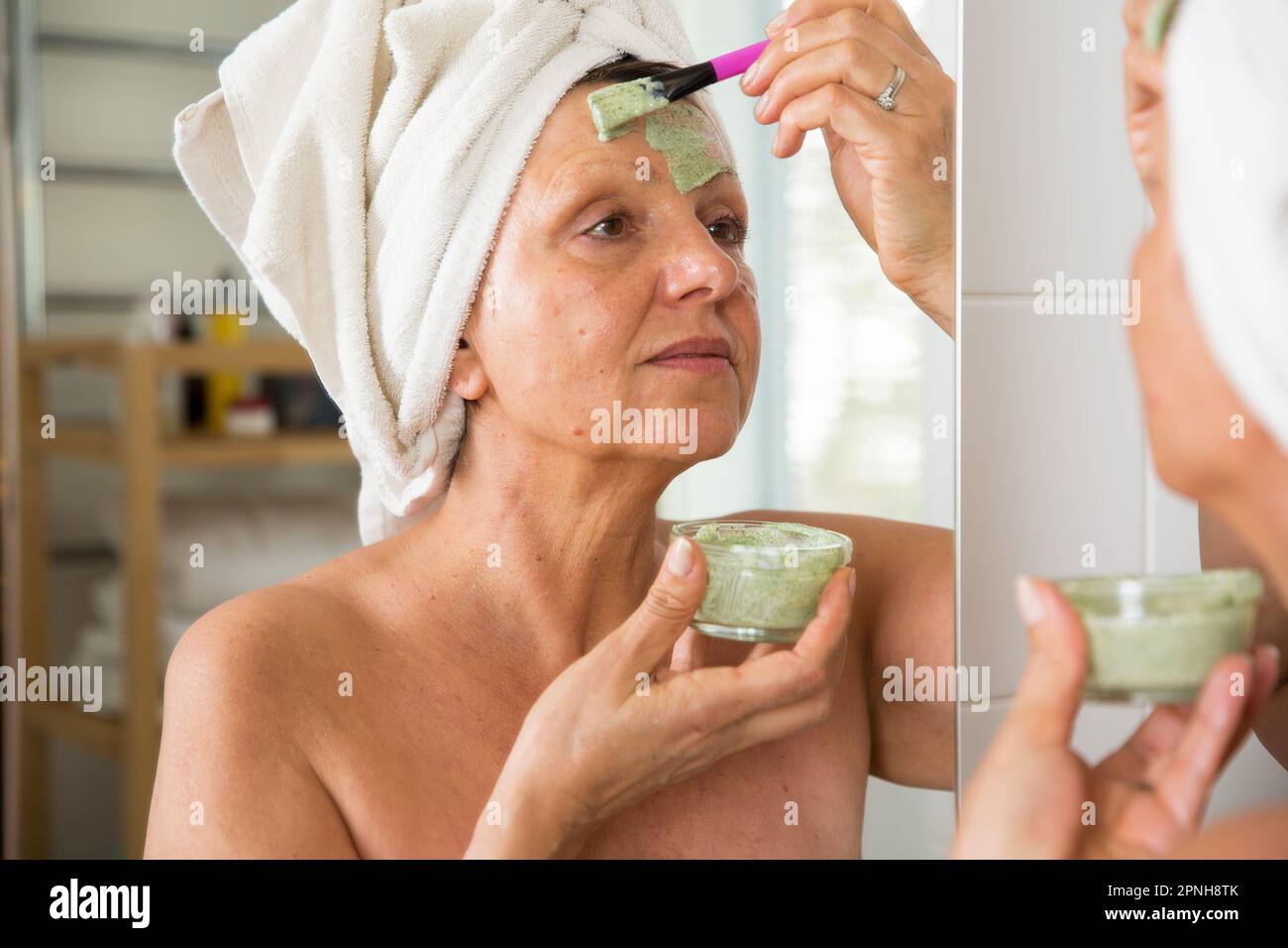 Reife, schöne Frau, die eine grüne hausgemachte Heimwerker-Gesichtsmaske auf der Haut aufträgt. In den Spiegel im Bad, in ein Handtuch gewickelt. Hautpflege-Routine Stockfoto