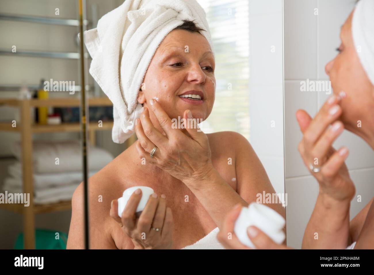 Reife, schöne Frau, die feuchtigkeitsspendende Gesichtscreme aus dem Weithalsgefäß auf die Haut aufträgt. In den Spiegel im Bad, in ein Handtuch gewickelt. Hautpflege-Routine Stockfoto