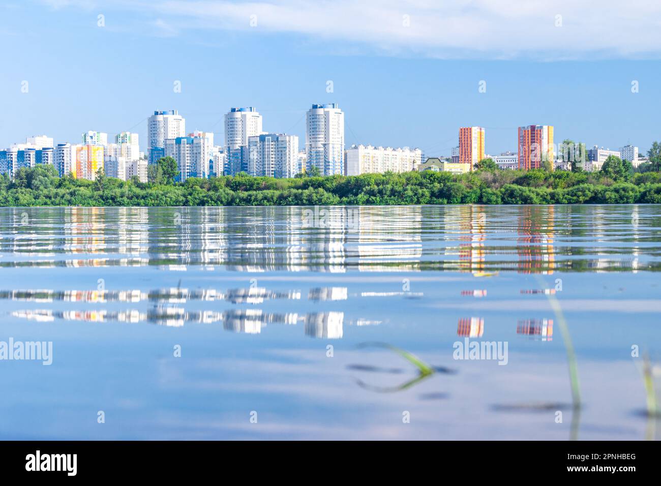Blau-weiße und rot-gelbe Hochhäuser hinter der Küstenvegetation spiegeln sich im Wasser eines Sees oder Flusses wider, selektiver Fokus Stockfoto