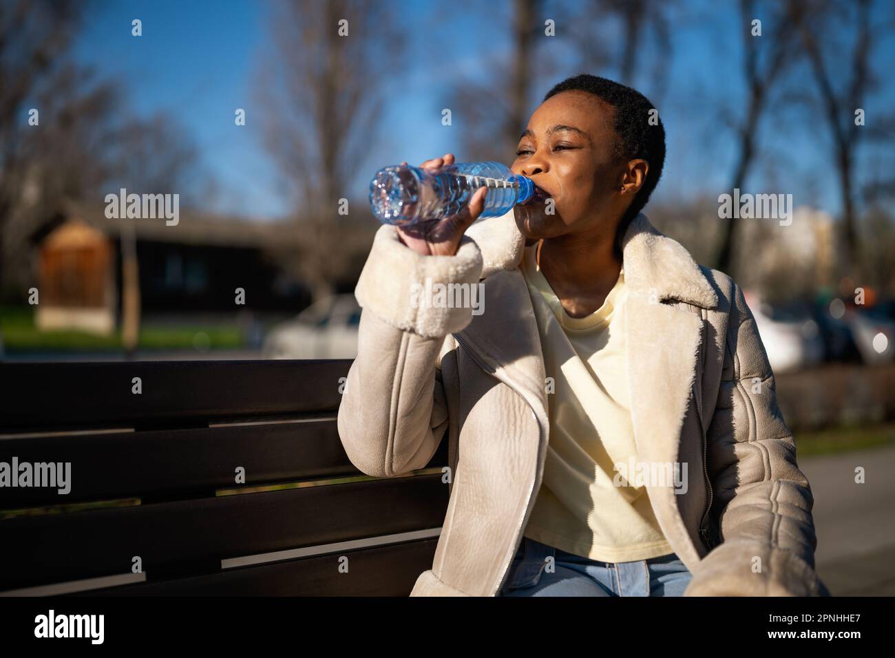 Außenporträt einer glücklichen afroamerikanischen Frau am sonnigen Tag. Sie sitzt auf einer Bank auf der Straße und trinkt Wasser. Stockfoto