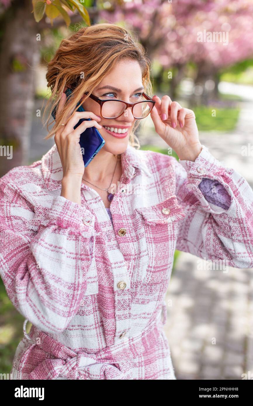 Glückliche junge, erfolgreiche Geschäftsfrau mit Brille, die im Frühling draußen anruft Stockfoto