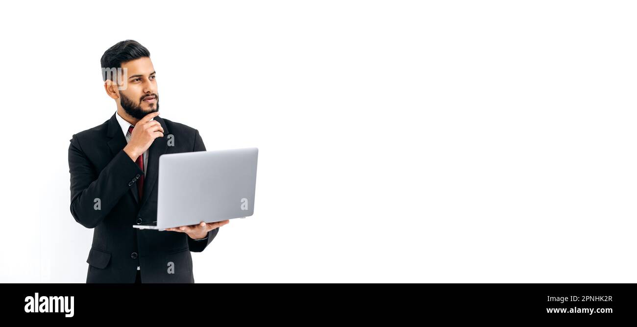 Panoramafoto eines verwirrten, durchdachten, eleganten arabers oder indianers im Anzug, auf isoliertem weißem Hintergrund mit Laptop stehen, nachdenklich wegschauen, träumen. Kopierraum für Ihre Präsentation Stockfoto