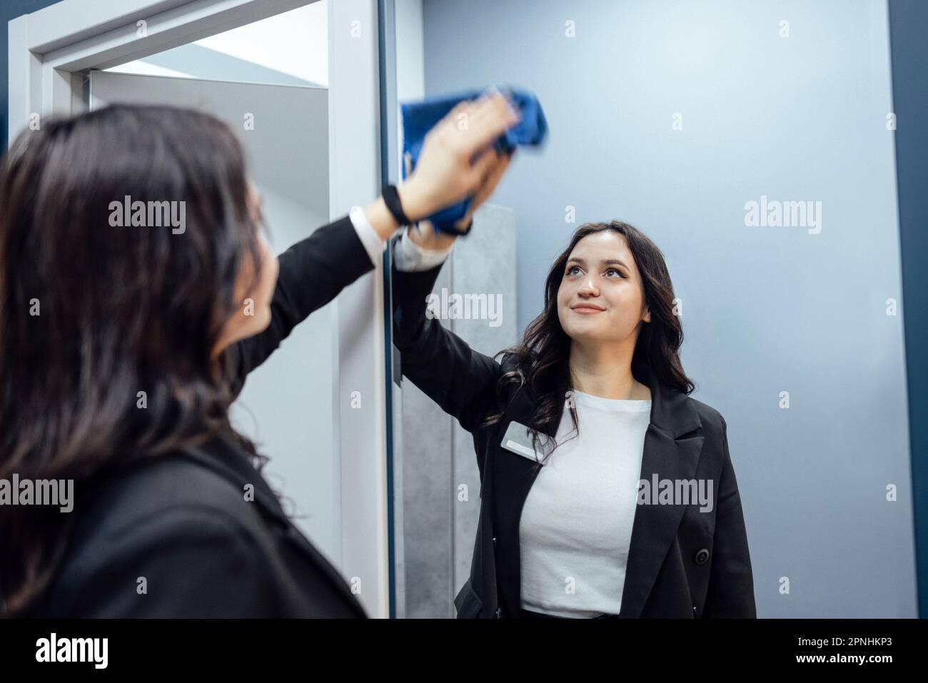 Junge brünette Frau in schwarzer Jacke wischt Spiegel im Fitnessclub. Das Personal räumt im Fitnessraum auf. Modernes, elegantes, helles Interieur mit Sportausrüstung, Stockfoto