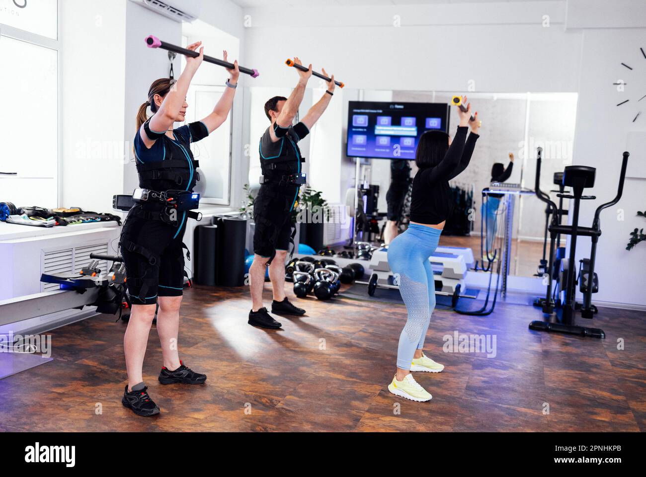 Sportpaar im rettungsdienst trainiert mit Personal Trainer im Fitnessclub. Junge Leute halten Sportstifte in der Hand und machen Squats. Der Coach zeigt das e Stockfoto