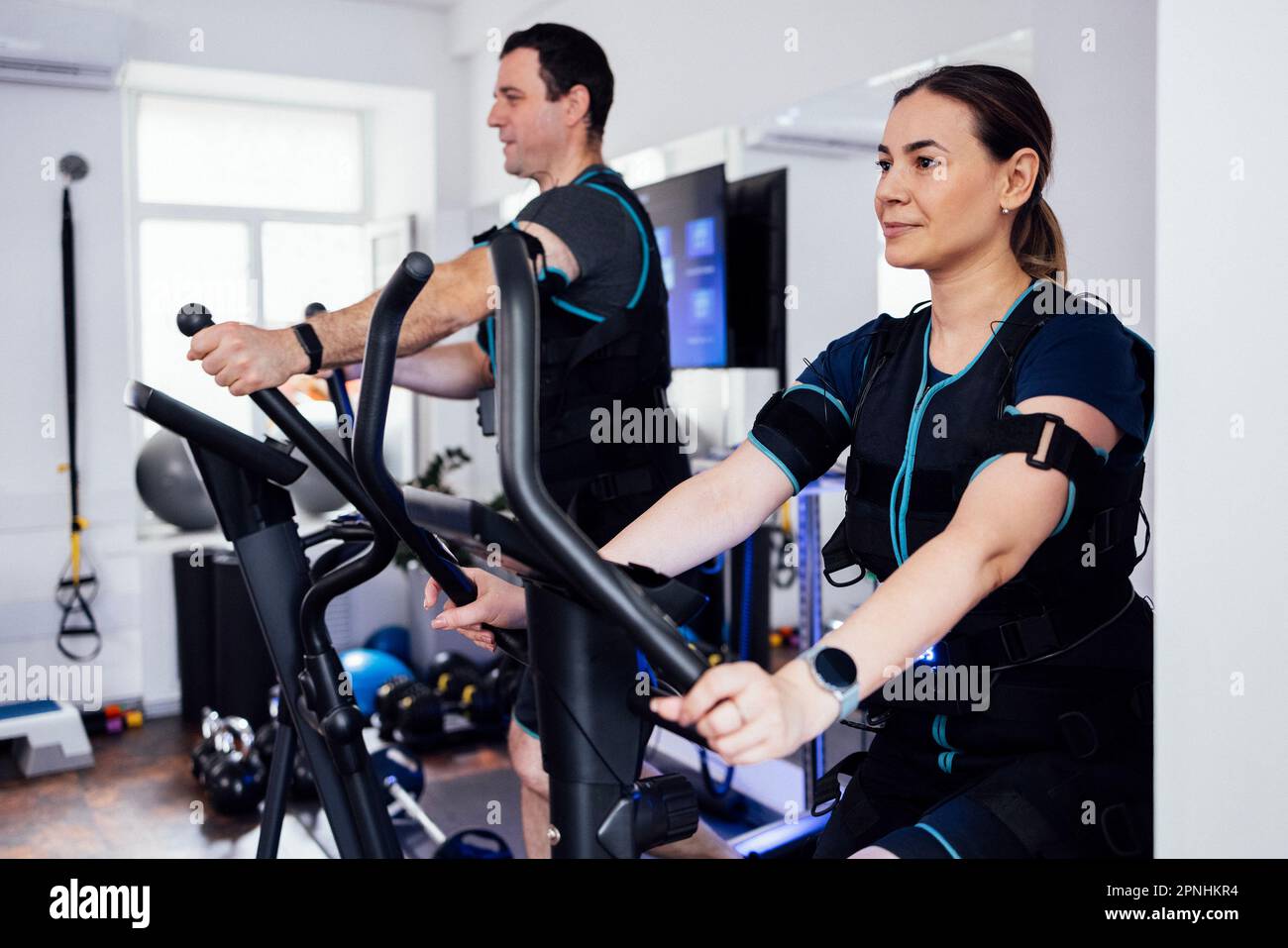 Einige Sportler in ems-Anzügen sind im Fitnessclub oder Fitnessraum mit Sportgeräten beschäftigt. Der lachende junge Mann trainiert auf dem Crosstrainer. P Stockfoto