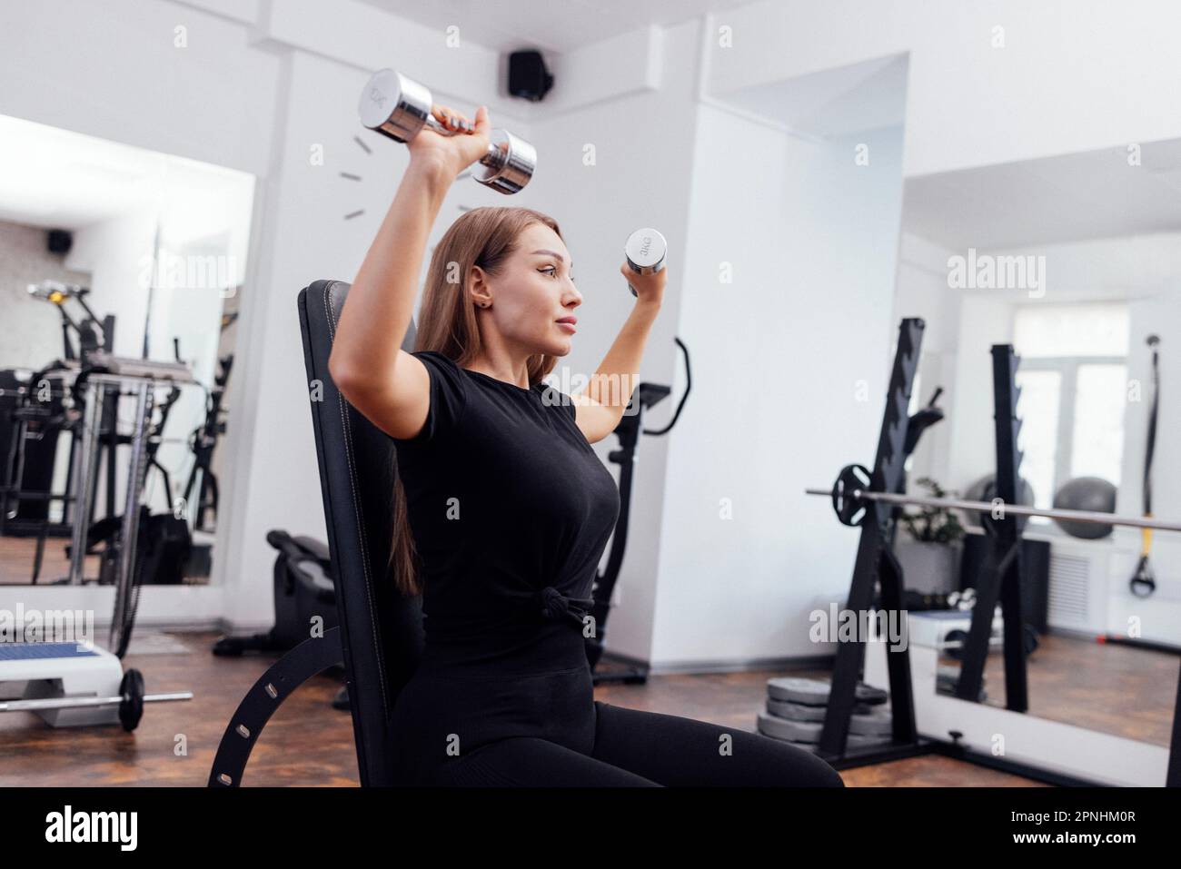 Eine sportliche junge blonde Frau in schwarzer Sportbekleidung presst auf einer geneigten Universalbank im Fitnessstudio oder Fitnessstudio die Kurzhantel. Pumpen der Muskeln von b Stockfoto