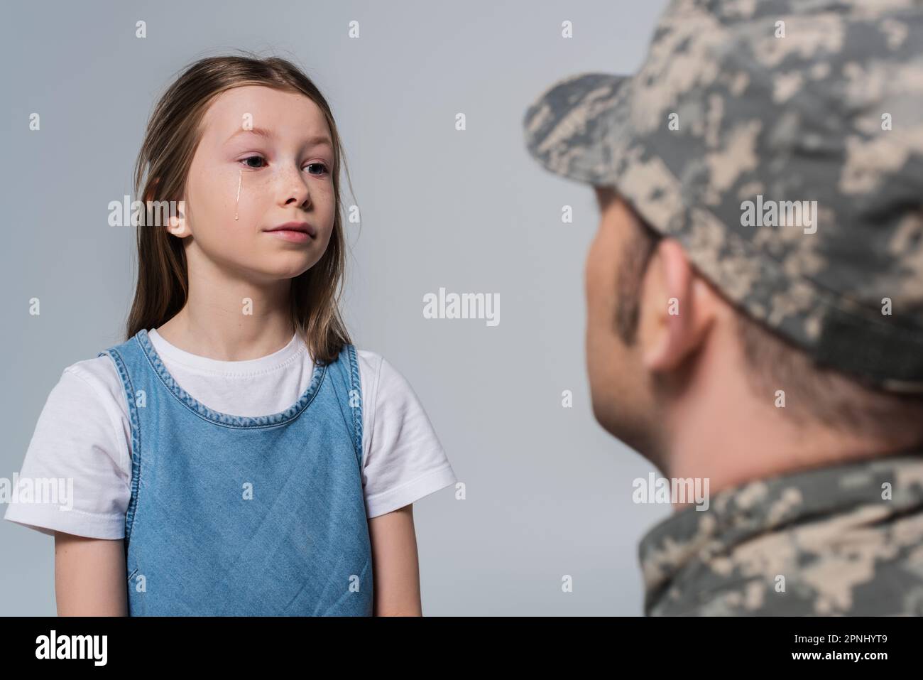 Ein junges Mädchen, das den tapferen Vater in Armeeuniform ansieht, auf verschwommenem Vordergrund isoliert auf Grau Stockfoto