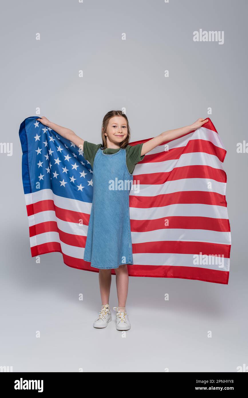 Die ganze Länge des patriotischen Mädchens, das lächelt, während es während des Gedenkfeiertags die riesige Flagge der Vereinigten Staaten auf grau hielt Stockfoto