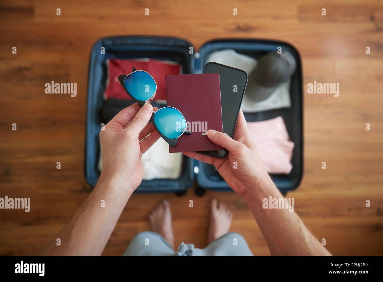Koffer packen und Sommerabenteuer planen. Ein Mann, der ein Smartphone, einen Reisepass und eine Sonnenbrille in der Hand über dem offenen Gepäck hält. Stockfoto