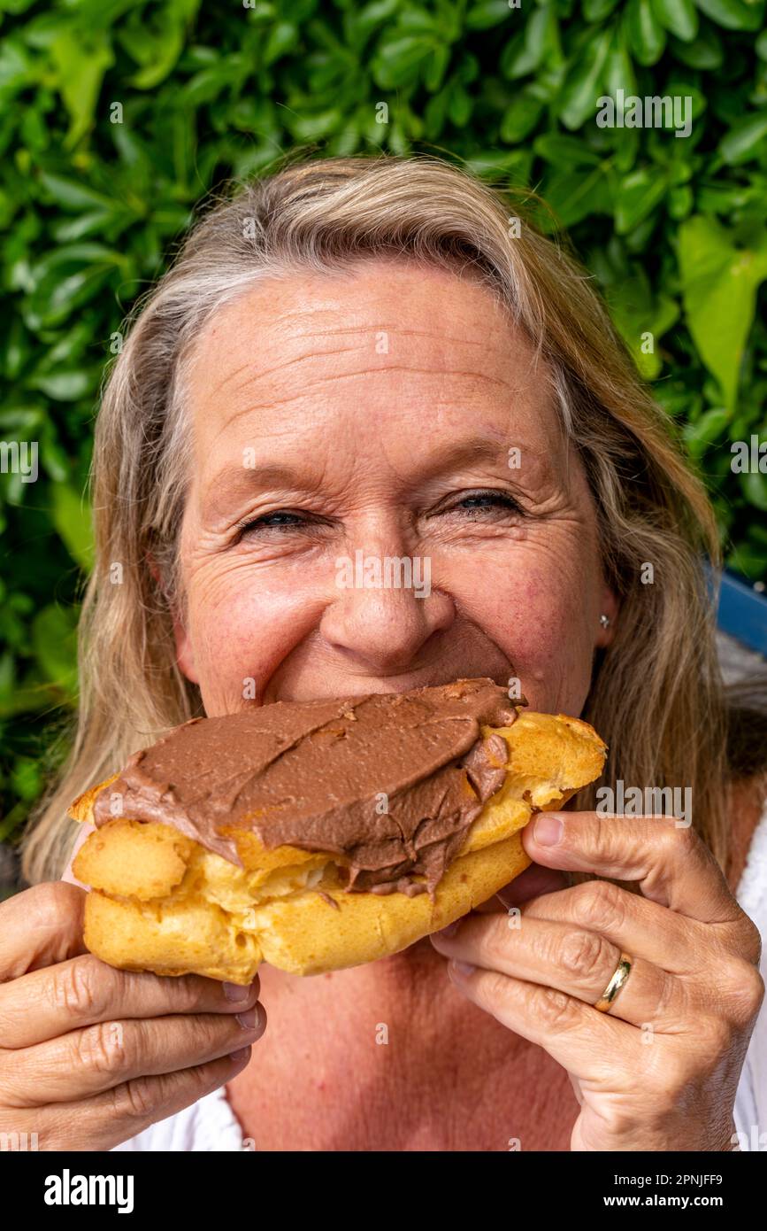 Eine Frau mittleren Alters, die Ein Chocolate Eclair isst, vor dem Chocolate Eclair Shop in Ohakune, North Island, Neuseeland Stockfoto
