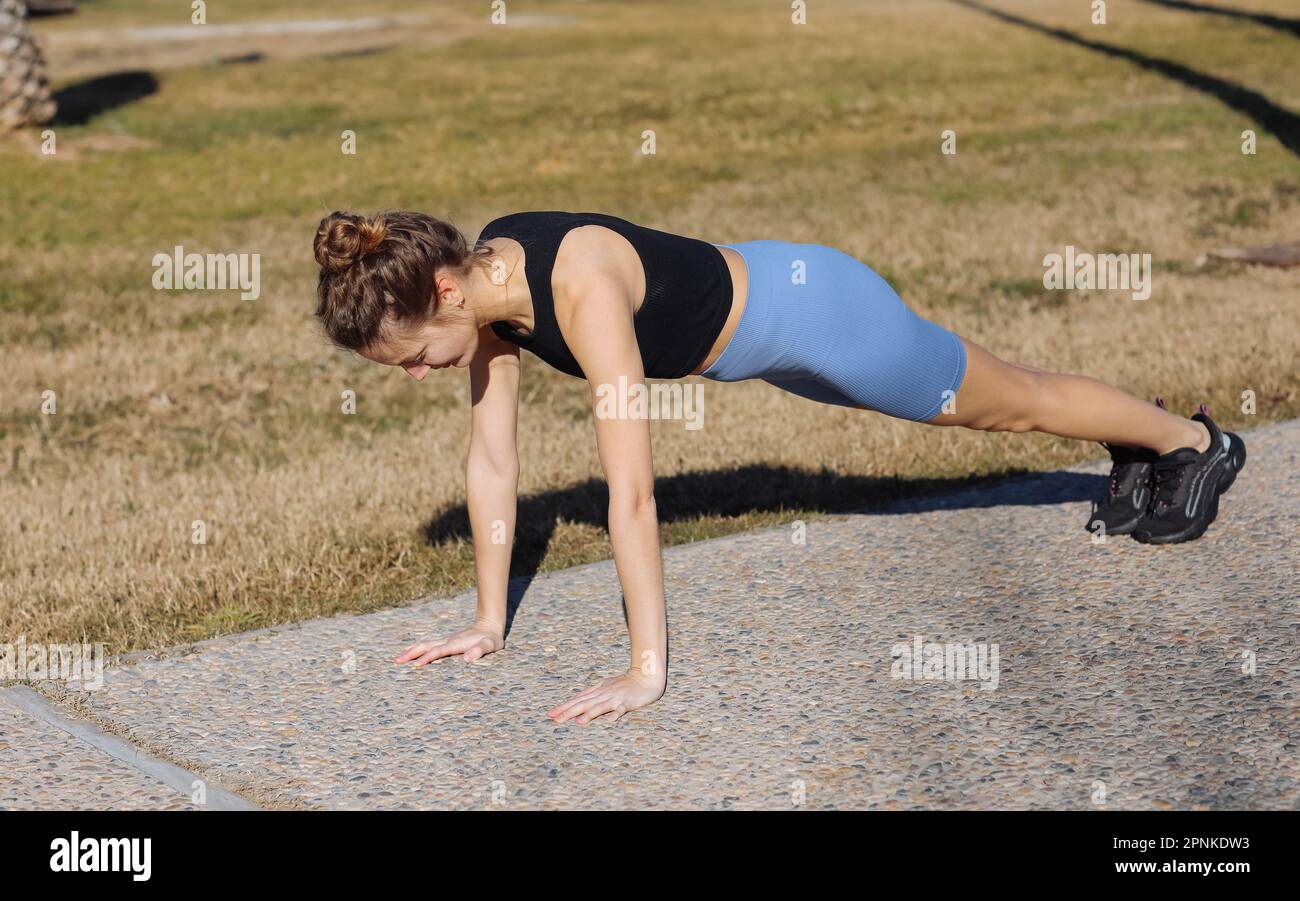 Weltgesundheitstag. Wunderschön fit, junge Frau in Sportbekleidung, trainiert draußen im Park, macht Stretching-Übungen in Pose, Hund, Maulkorb, Sonne Stockfoto