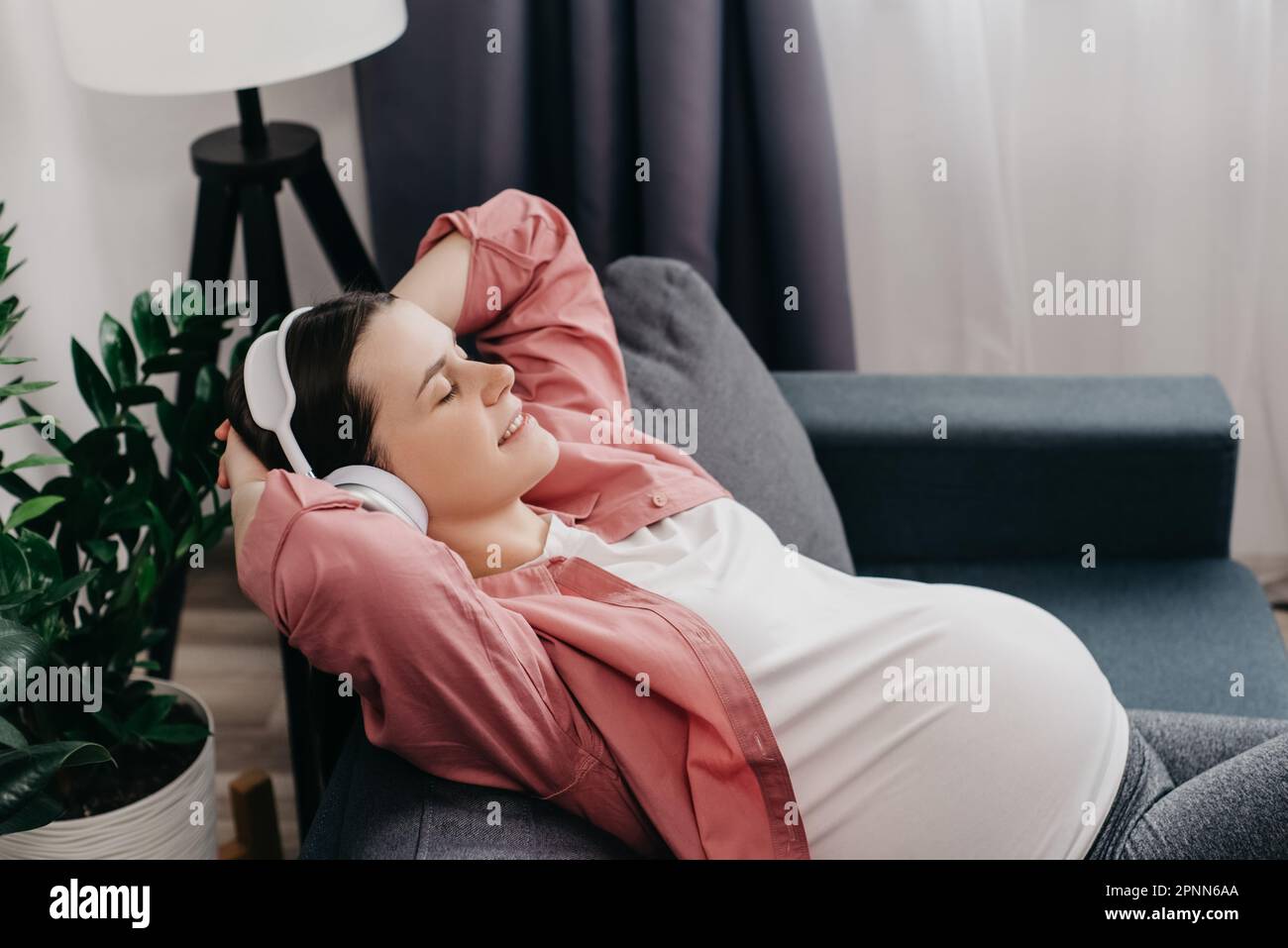 Lächelnde junge Schwangere, die sich auf dem Sofa lehnt und Kopfhörer trägt, die Augen schließt und Musik hört. Genießen Sie den Audiotitel Ihres Lieblingssongs. Freizeit Stockfoto