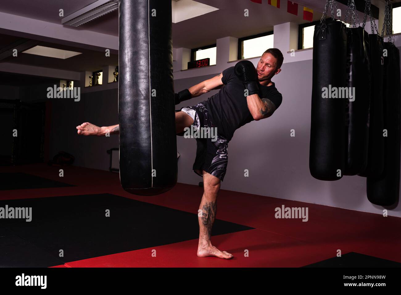 Ein weißer Mann, der ein schwarzes T-Shirt und Shorts trägt und eine hängende Boxtasche im Fitnessstudio schlägt Stockfoto